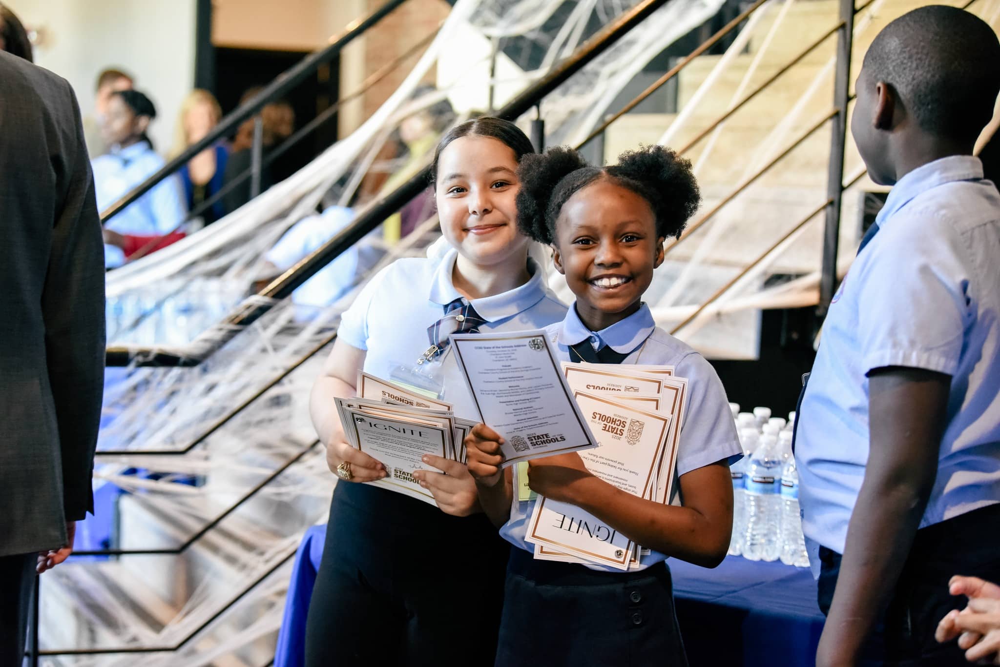 Two students in uniforms smiling and holding IGNITE certificates at a school recognition ceremony.